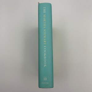 Puede incluir: Lomo de libro de cocina turquesa con letras doradas que dice "THE MARTHA STEWART COOKBOOK". La editorial, "POTTER", est&aacute; impresa en la parte inferior. El libro est&aacute; sobre un fondo blanco liso.