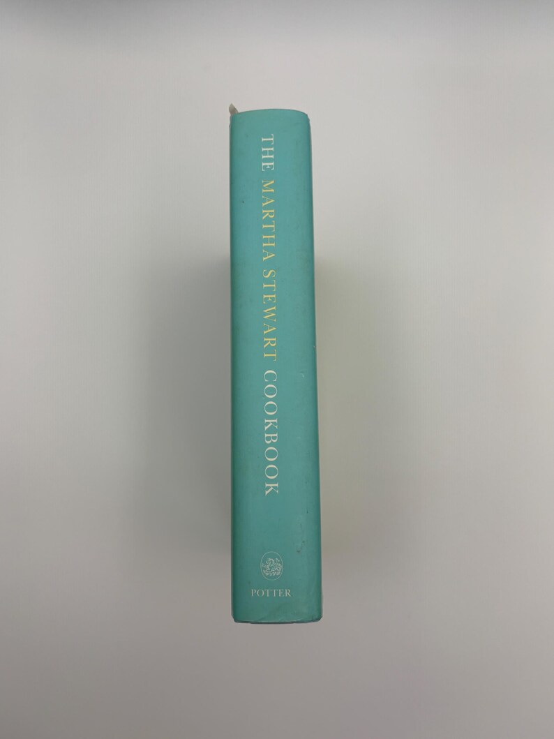 Puede incluir: Lomo de libro de cocina turquesa con letras doradas que dice "THE MARTHA STEWART COOKBOOK". La editorial, "POTTER", est&aacute; impresa en la parte inferior. El libro est&aacute; sobre un fondo blanco liso.