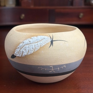 May include: A beige ceramic bowl with a feather design. The bowl has a rounded shape with a gray and black band around the middle. A white and gray feather is painted on the side.