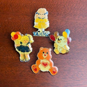 May include: A collection of vintage enamel pins featuring cartoon characters. The pins include a girl named Tammy, a teddy bear with a heart, and other characters holding balloons. The pins are in various colors, including yellow, orange, and blue.