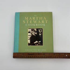Puede incluir: Un libro de cocina de tapa dura titulado "The Martha Stewart Cookbook" con una cubierta verde y una foto de Martha Stewart. El libro tiene un lomo turquesa y una regla de madera colocada en la parte inferior.