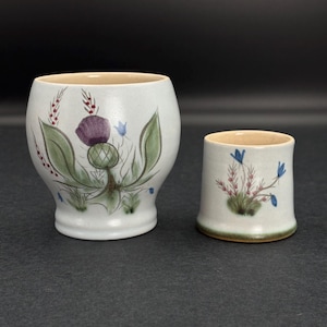 May include: Two ceramic vessels with floral designs. The larger, bulbous pot features a purple thistle, green leaves, and small red and blue accents. The smaller, cylindrical cup has a similar floral motif. Both have a cream-colored interior.