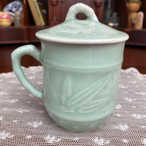 May include: A celadon green ceramic mug with a matching lid. The mug has a handle and a bamboo leaf design. The lid has a small loop for easy removal. The mug is sitting on a white lace tablecloth.
