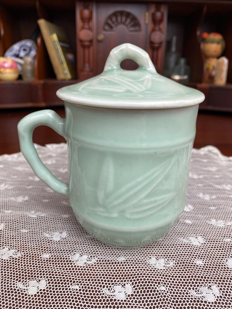 May include: A celadon green ceramic mug with a matching lid. The mug has a handle and a bamboo leaf design. The lid has a small loop for easy removal. The mug is sitting on a white lace tablecloth.