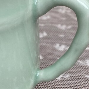 May include: Close-up of a pale green ceramic mug with a curved handle. The handle is smooth and glossy, and the mug has a rounded shape. The mug is likely handmade and has a unique, organic feel.
