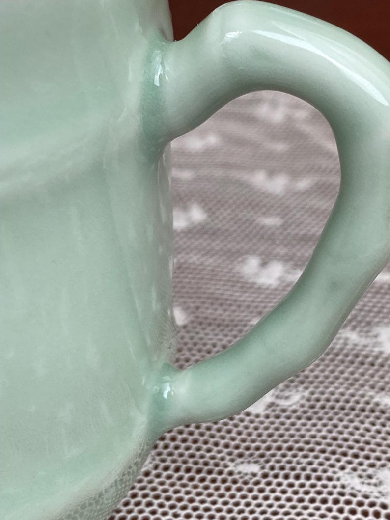 May include: Close-up of a pale green ceramic mug with a curved handle. The handle is smooth and glossy, and the mug has a rounded shape. The mug is likely handmade and has a unique, organic feel.