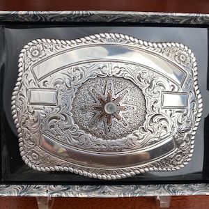 May include: Silver-toned rectangular belt buckle with intricate floral and scrollwork designs. The center features a starburst design with a small, clear stone. The buckle is presented in a black display box.