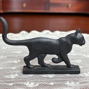 May include: A black cast iron cat figurine. The cat is depicted in a walking pose with its tail curved upwards. The figurine is mounted on a rectangular base. The cat statue is a decorative item.