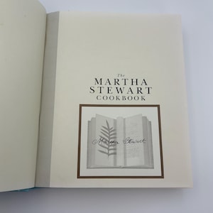 Puede incluir: Libro de cocina abierto titulado "The Martha Stewart Cookbook". La portada presenta el t&iacute;tulo en una fuente serif por encima de una ilustraci&oacute;n enmarcada de un libro abierto con un helecho y la firma del autor. Las p&aacute;ginas son de color blanquecino.