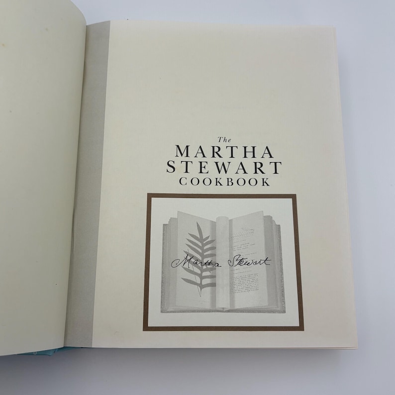 Puede incluir: Libro de cocina abierto titulado "The Martha Stewart Cookbook". La portada presenta el t&iacute;tulo en una fuente serif por encima de una ilustraci&oacute;n enmarcada de un libro abierto con un helecho y la firma del autor. Las p&aacute;ginas son de color blanquecino.