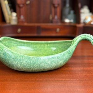 May include: A green ceramic serving dish with a curved handle. The dish has a speckled texture and a glossy finish. The handle curves upward and ends in a rounded tip. The dish is on a wooden surface.