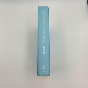 Puede incluir: Un libro de cocina azul claro titulado "The Martha Stewart Cookbook" con letras doradas. El lomo del libro presenta el logotipo de Potter en la parte inferior. El libro est&aacute; de pie sobre un fondo blanco.