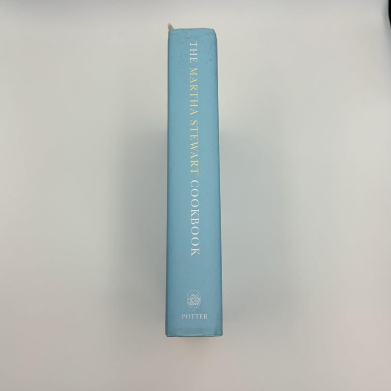 Puede incluir: Un libro de cocina azul claro titulado "The Martha Stewart Cookbook" con letras doradas. El lomo del libro presenta el logotipo de Potter en la parte inferior. El libro est&aacute; de pie sobre un fondo blanco.