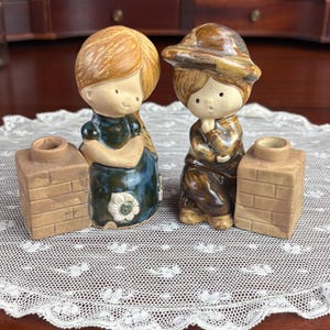 May include: Two ceramic figurines of children, one with long hair and a blue dress with a white flower, the other wearing a brown hat and outfit. Each figure is positioned next to a small, square, tan-colored ceramic chimney-shaped candle holder.