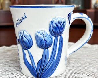 May include: White ceramic mug with blue tulip design and handle. The word "Holland" is written in blue script on the side. The mug has a blue rim and blue accents on the handle. The mug is sitting on a white lace tablecloth.