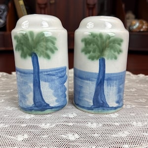 May include: A pair of ceramic salt and pepper shakers featuring a hand-painted design of a green palm tree against a blue ocean background. The shakers have a cylindrical shape with a rounded top and a smooth, off-white glazed finish.