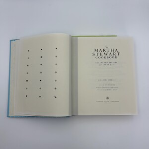 Puede incluir: Un libro de cocina abierto titulado "The Martha Stewart Cookbook" con una cubierta azul claro. La p&aacute;gina izquierda presenta una cuadr&iacute;cula de s&iacute;mbolos negros, mientras que la derecha muestra el t&iacute;tulo y el nombre del autor.
