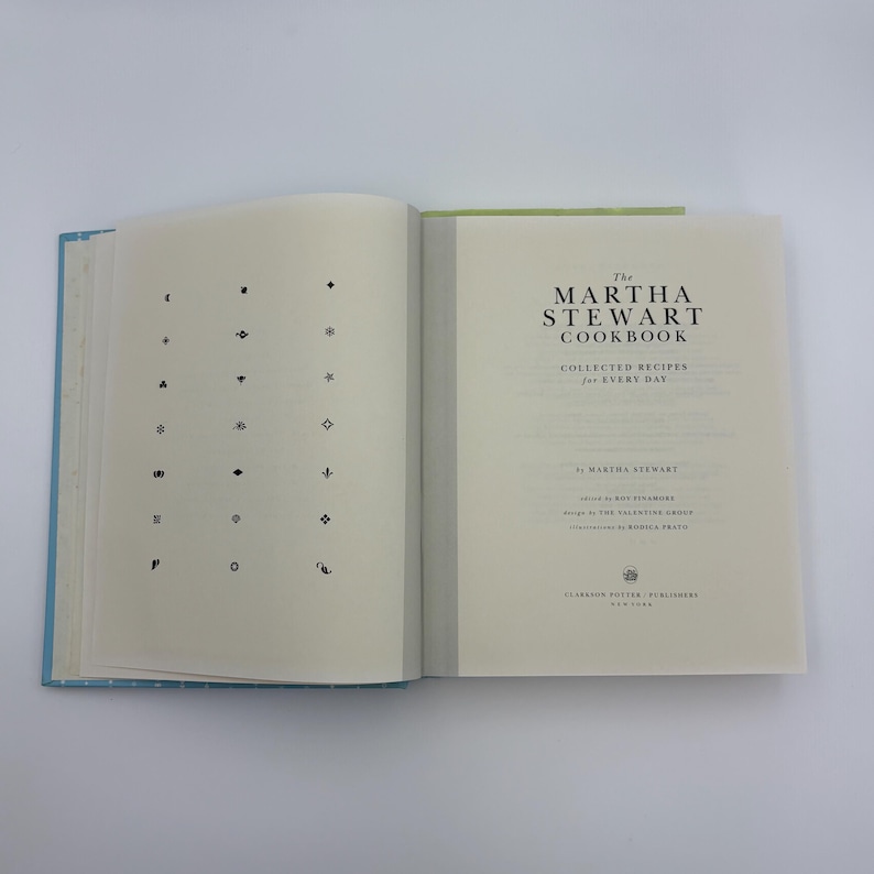 Puede incluir: Un libro de cocina abierto titulado "The Martha Stewart Cookbook" con una cubierta azul claro. La p&aacute;gina izquierda presenta una cuadr&iacute;cula de s&iacute;mbolos negros, mientras que la derecha muestra el t&iacute;tulo y el nombre del autor.