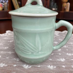 May include: A pale green ceramic mug with a matching lid. The mug features a handle and a bamboo leaf design. The lid has a small loop. The mug is approximately 10 cm tall.