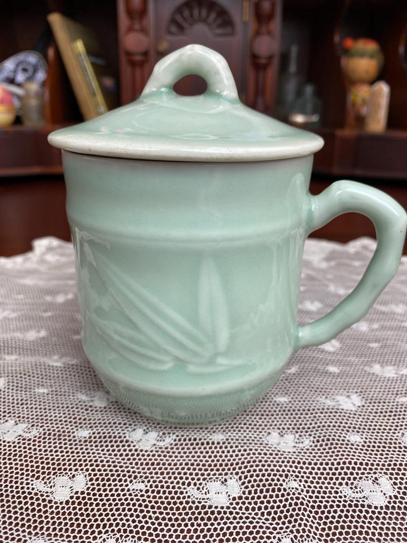 May include: A pale green ceramic mug with a matching lid. The mug features a handle and a bamboo leaf design. The lid has a small loop. The mug is approximately 10 cm tall.