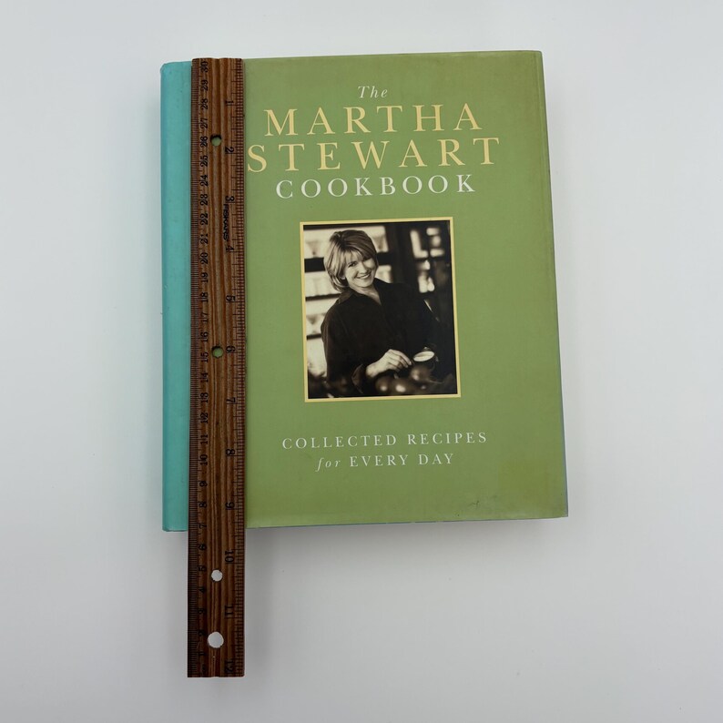 Puede incluir: Un libro de cocina de tapa dura titulado "The Martha Stewart Cookbook" con una cubierta verde y una foto de Martha Stewart. El libro tiene un lomo turquesa y una regla de madera colocada junto a &eacute;l. El texto "COLLECTED RECIPES for EVERY DAY" est&aacute; impreso en la portada.
