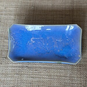 May include: Rectangular ceramic serving dish with a glossy, blue glaze. The dish features a leaf-like pattern and a light green trim. The edges are slightly curved, and the overall shape is rectangular.
