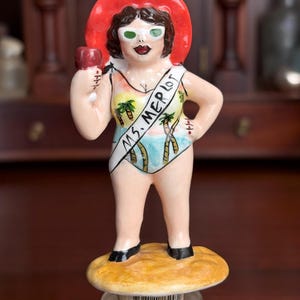 May include: A ceramic bottle stopper featuring a figurine of a woman in a swimsuit and a red sun hat. The figurine holds a red object and wears a sash that reads "MS. MERLOT". The swimsuit has a tropical design. The stopper is on a bottle.