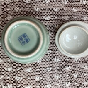 May include: A celadon ceramic teacup and lid. The cup has a small handle and a blue stamp on the base. The lid is round and is next to the cup. The set is on a white lace surface.
