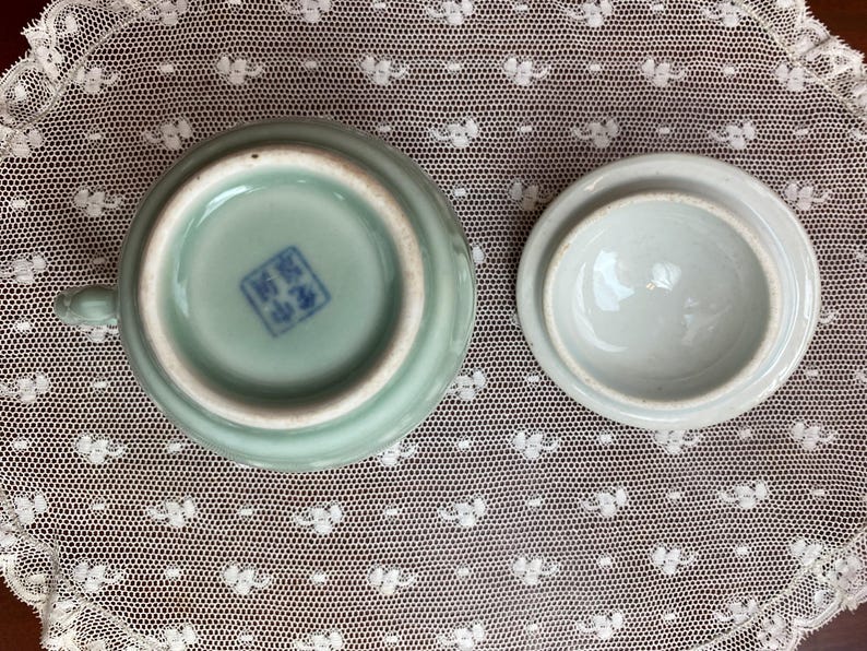 May include: A celadon ceramic teacup and lid. The cup has a small handle and a blue stamp on the base. The lid is round and is next to the cup. The set is on a white lace surface.