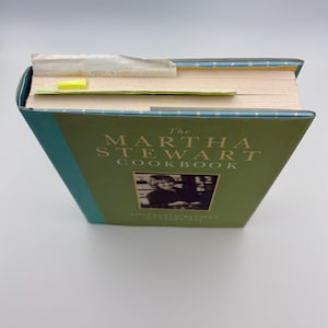 Puede incluir: Un libro de cocina de tapa dura titulado "The Martha Stewart Cookbook" con una cubierta verde y lomo turquesa. La portada presenta el t&iacute;tulo en letras blancas y una foto en blanco y negro. Una nota adhesiva amarilla se utiliza como marcador.