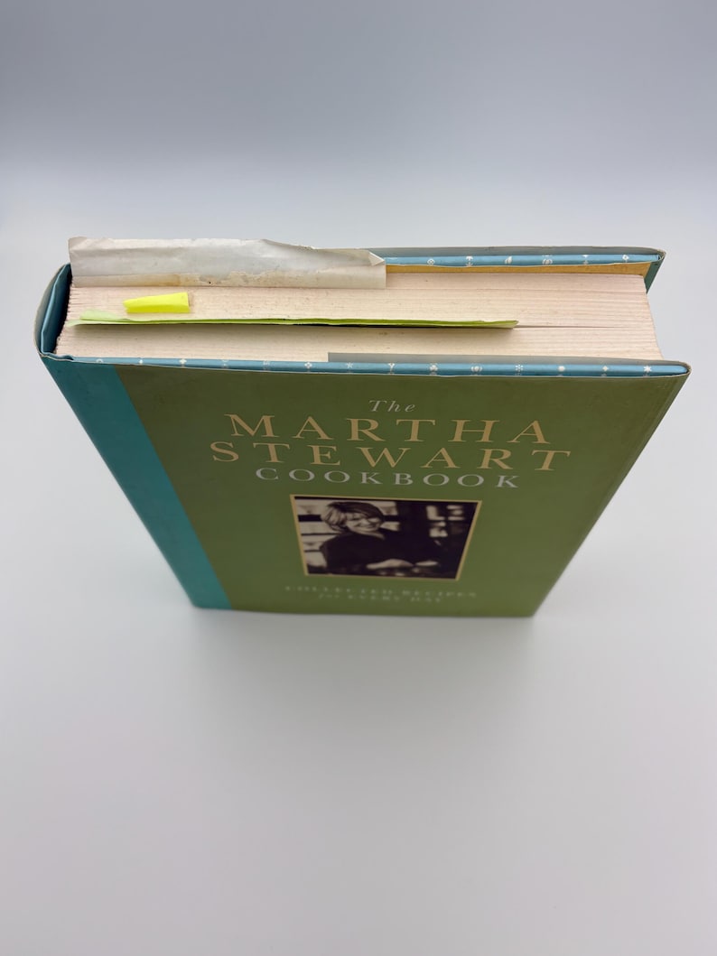 Puede incluir: Un libro de cocina de tapa dura titulado "The Martha Stewart Cookbook" con una cubierta verde y lomo turquesa. La portada presenta el t&iacute;tulo en letras blancas y una foto en blanco y negro. Una nota adhesiva amarilla se utiliza como marcador.