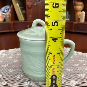 May include: A celadon green ceramic mug with a lid, featuring a handle and bamboo leaf design. The mug is approximately 10 cm tall, as measured by a yellow tape measure.