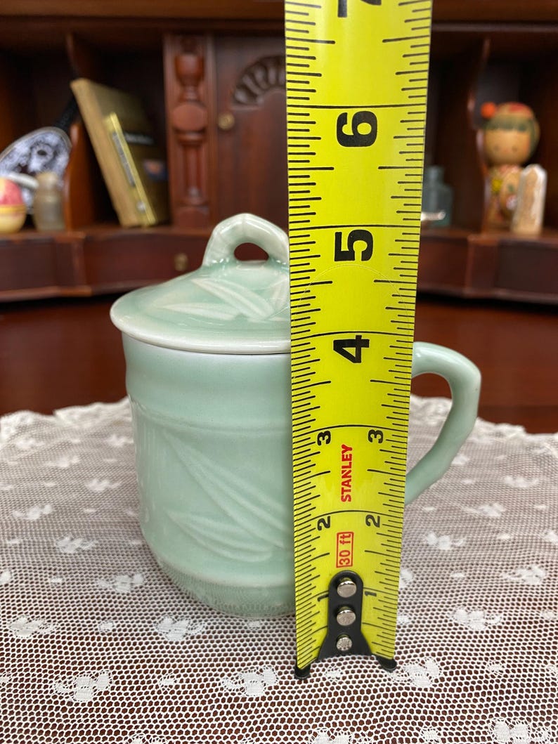 May include: A celadon green ceramic mug with a lid, featuring a handle and bamboo leaf design. The mug is approximately 10 cm tall, as measured by a yellow tape measure.