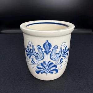 May include: A white ceramic utensil holder with a blue floral and bird design. The cylindrical holder has a slightly flared rim and a blue band around the top. The design features two birds facing each other with floral accents.