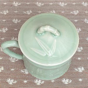 May include: A pale green ceramic mug with a matching lid. The lid has a bamboo-shaped handle and bamboo leaf designs. The mug has a handle and a textured pattern around the base. The mug is on a white lace doily.