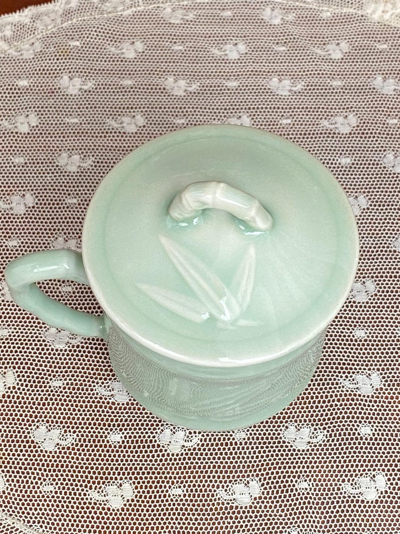 May include: A pale green ceramic mug with a matching lid. The lid has a bamboo-shaped handle and bamboo leaf designs. The mug has a handle and a textured pattern around the base. The mug is on a white lace doily.