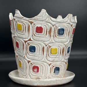 May include: A white ceramic planter with a matching saucer. The planter features a repeating pattern of squares in red, yellow, and blue, outlined in brown. The rim has a scalloped edge. The planter is approximately 7 inches tall and 8 inches wide.