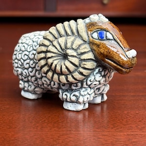 May include: A ceramic ram figurine with a detailed design. The ram has a tan head and horns, with a blue eye. The body is white with a textured pattern. The figurine is a decorative item.