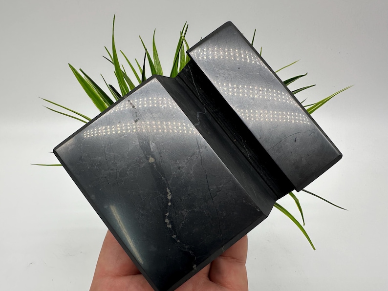 May include: A dark gray, geometric, angled sculpture with a textured surface. The sculpture is made of a solid material and has a folded, zig-zag design. Green grass-like plants are behind the sculpture.