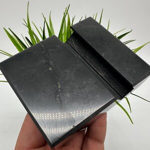 May include: A black, rectangular phone stand made of stone. The stand has a slot to hold a phone and is angled for easy viewing. Green plants are visible behind the stand.