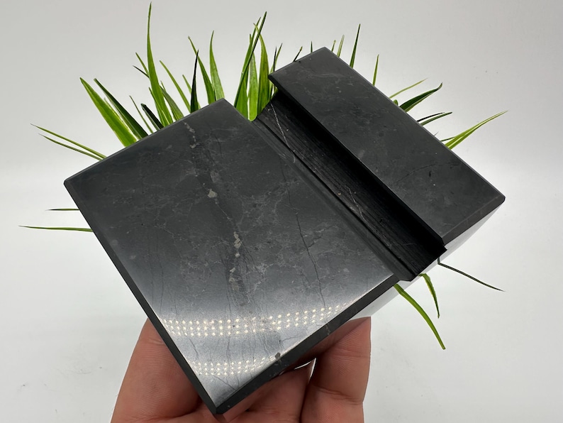 May include: A black, rectangular phone stand made of stone. The stand has a slot to hold a phone and is angled for easy viewing. Green plants are visible behind the stand.
