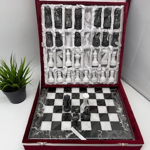 Könnte beinhalten: Ein Schachspiel mit einem schwarz-weißen Marmorbrett und passenden Steinen. Das Set befindet sich in einem mit tiefrotem Samt ausgekleideten Koffer mit einzelnen Fächern für die Figuren. Vier Schachfiguren sind auf dem Brett aufgestellt.