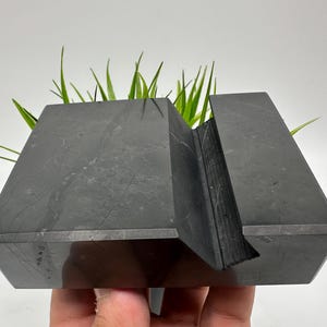 May include: A dark gray, geometric stone planter with a slot for holding a phone or tablet. The planter has a modern, angular design and is filled with green grass-like plants. The planter is held by a hand.