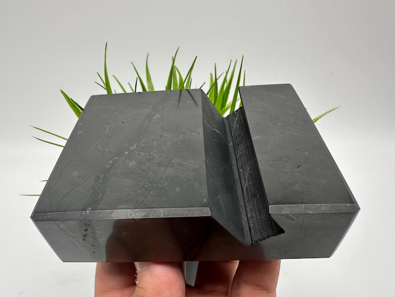 May include: A dark gray, geometric stone planter with a slot for holding a phone or tablet. The planter has a modern, angular design and is filled with green grass-like plants. The planter is held by a hand.