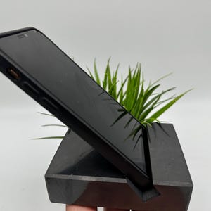 May include: A black smartphone rests on a black, angled phone stand. The stand has a slot to hold the phone upright. Green plant leaves are visible behind the phone and stand. The stand is made of a dark, solid material.