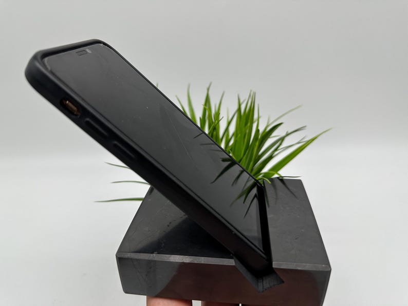 May include: A black smartphone rests on a black, angled phone stand. The stand has a slot to hold the phone upright. Green plant leaves are visible behind the phone and stand. The stand is made of a dark, solid material.