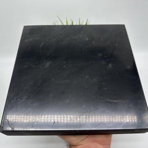 May include: A black square slate tile with a smooth, polished surface. The tile has a subtle sheen and a slightly textured appearance.