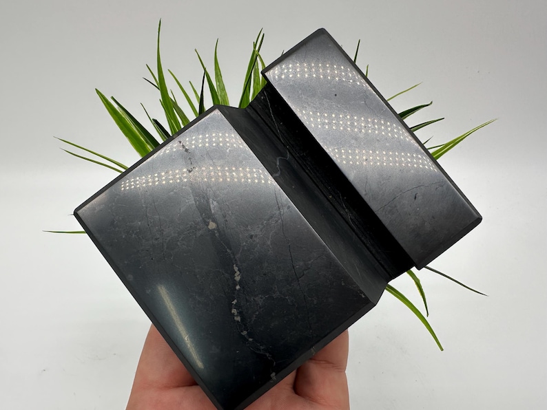 May include: A black, geometric, folded stone sculpture with a reflective surface. The sculpture is a dark gray color and has a textured appearance. Green grass-like plants are behind the sculpture.