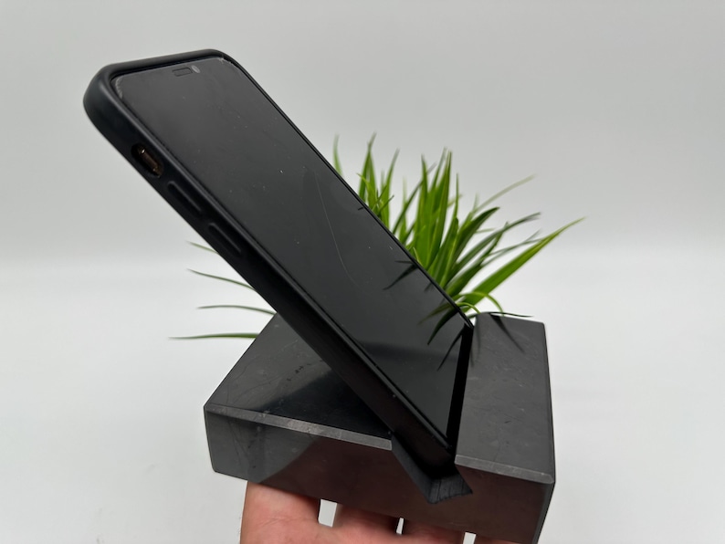 May include: A black smartphone in a black case is propped up on a dark gray, angled phone stand. The stand has a slot to hold the phone and is made of a solid, rectangular block. Green plants are in the background.