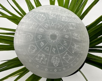 May include: A white selenite charging plate with a zodiac wheel design etched into the surface. The wheel features the twelve zodiac signs and symbols, including Aries, Taurus, Gemini, Cancer, Leo, Virgo, Libra, Scorpio, Sagittarius, Capricorn, Aquarius, and Pisces.